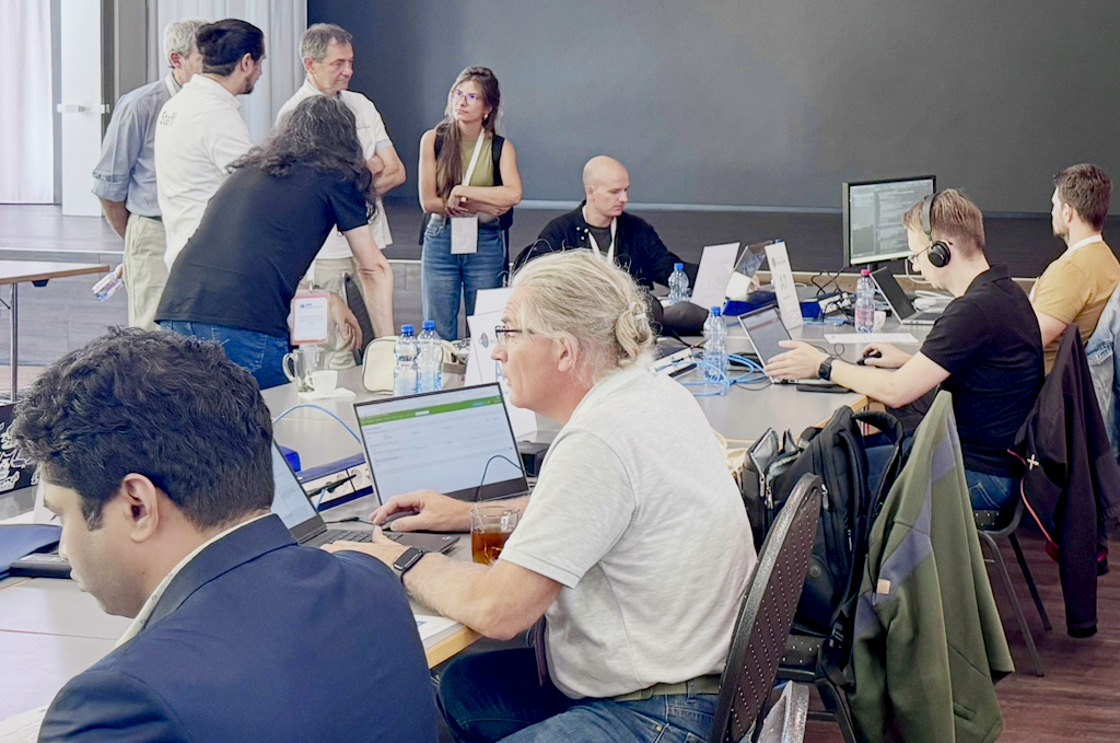 Bild von der Führung während des Projectathon 2025. Man sieht Leute, die an ihren PCs arbeiten, und ein paar Leute, die daneben stehen und denen erklärt wird, was die Experten gerade testen.